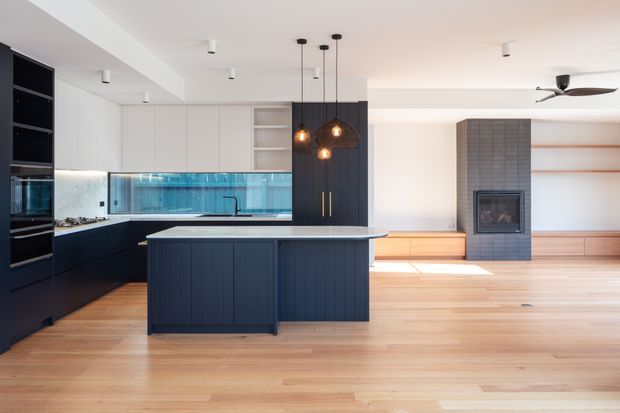Open-plan contemporary kitchen and living area with a dark navy island and cabinetry, pale timber flooring, white upper cabinetry, and a turquoise glass splashback. Warm pendant lights hang above the island. The adjoining living zone features a tiled fireplace surround, timber built-in joinery, recessed shelving, and a ceiling fan.