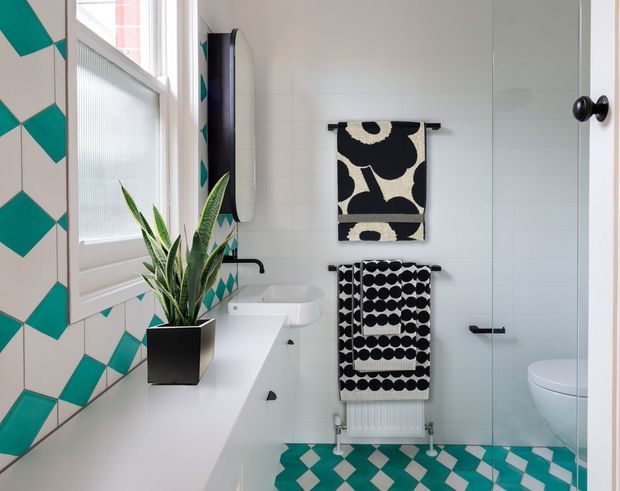 A bright, compact bathroom with white tiled walls, a white vanity and basin, a glass shower enclosure, and a partially visible toilet. The space features bold teal-and-white geometric floor and wall tiles, black-and-white patterned towels, a potted snake plant, and black tapware and hardware. A window provides natural light.