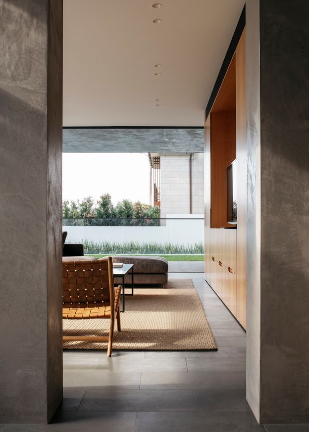 A refined contemporary living room viewed through a wide doorway, featuring smooth grey concrete-like walls, large-format grey floor tiles, a warm timber built-in media wall, and a neutral woven rug. A leather lounge chair and sofa face the built-in television, while full-height glazing opens toward a landscaped outdoor area and pool edge. Recessed ceiling lights and minimal detailing create a calm, architectural atmosphere.