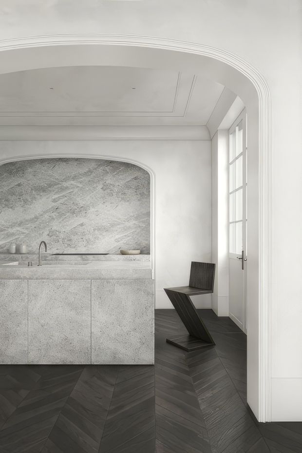 A refined monochromatic kitchen interior with a sculptural stone island and integrated sink, framed by a large rounded arch opening with a textured stone backsplash. Dark herringbone timber flooring contrasts with white plaster walls and an ornate curved ceiling detail. A tall glazed door provides natural light, while a minimalist sculptural chair sits beside the island.