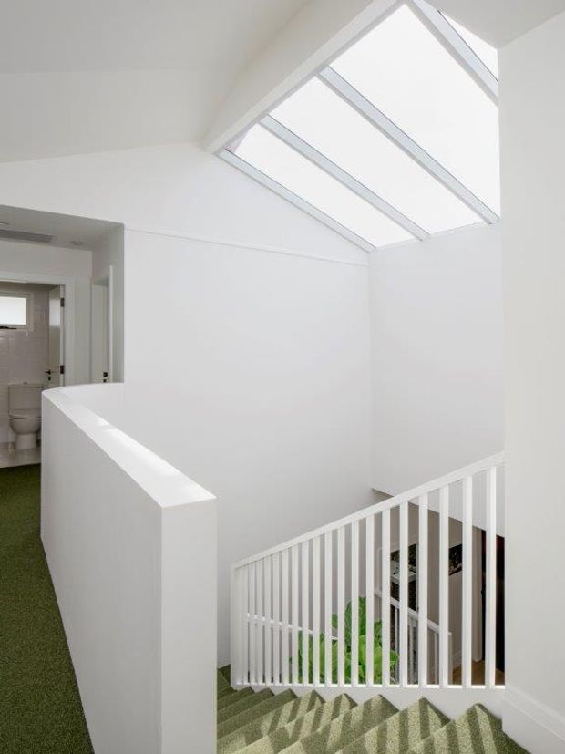 Bright white stairway and landing with a green carpeted staircase, white balustrade, and a large sloping skylight flooding the stairwell with natural light. A bathroom is visible through the doorway at left.