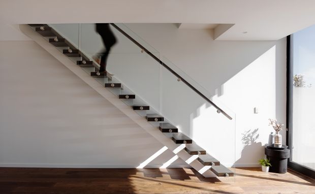 Bright contemporary stairway with open-riser timber treads, a slim dark handrail, and clear glass balustrades. The staircase is set against white walls and a white ceiling, with dark timber flooring and strong sunlight casting geometric shadows. A black cylindrical planter and small decorative plants sit beside the full-height glazing.