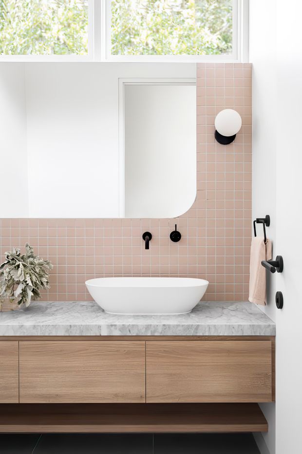 A bright contemporary bathroom vanity featuring a floating light-wood timber cabinet, grey marble-look stone benchtop, white vessel basin, large frameless mirror, and a blush-pink square tile splashback. Matte black wall-mounted tapware, towel rail, and circular wall light provide contrasting accents. A high window brings in abundant natural light.