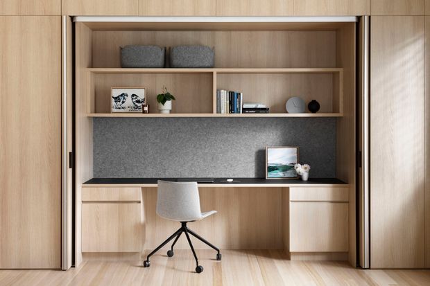 A compact built-in home office nook integrated into warm light-wood cabinetry. The desk is set beneath open shelving with books, decorative objects and plants, backed by a dark grey felt or fabric panel. A light upholstered office chair sits on pale timber flooring, and full-height timber doors frame the workspace.