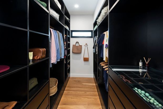 A compact, highly organised walk-in wardrobe with floor-to-ceiling black built-in shelving and hanging rails on both sides. The space has warm timber flooring, white walls, a small horizontal window, integrated drawers, open display shelving, handbags and clothing, and a glass-topped drawer unit displaying accessories.