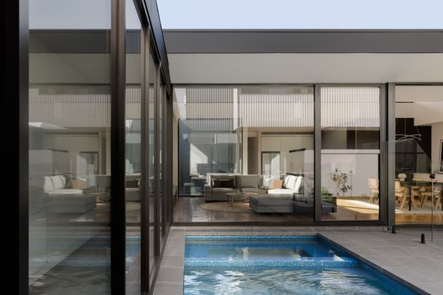 Contemporary courtyard interior view with extensive dark-framed floor-to-ceiling glazing surrounding a tiled swimming pool. A lounge area and dining space are visible through the glass, with strong indoor-outdoor connection and a clean, restrained material palette.