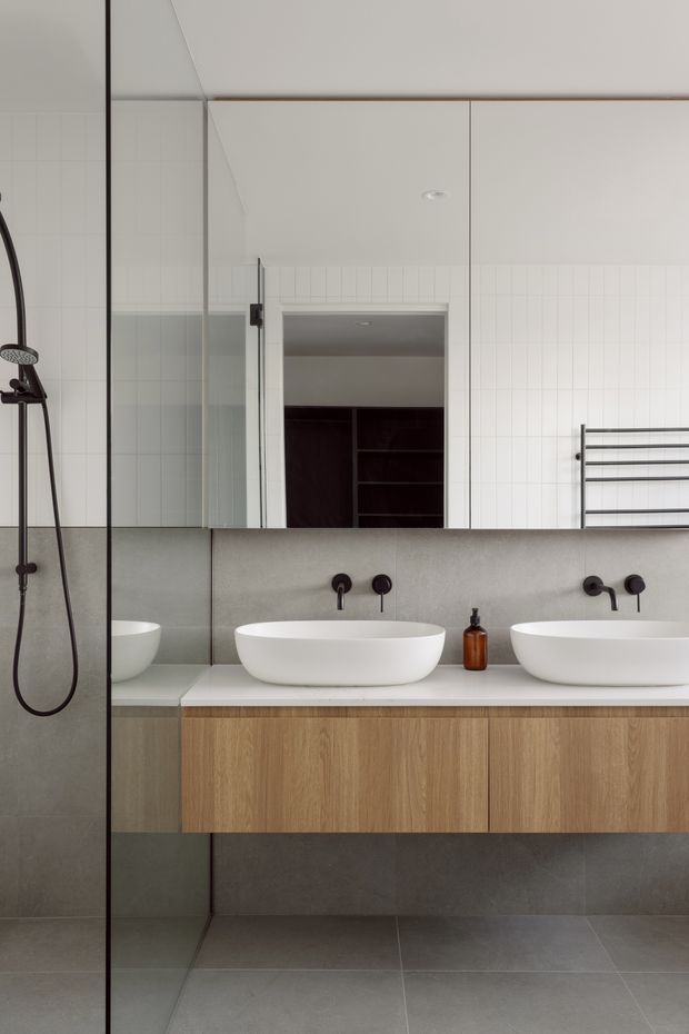 Contemporary bathroom with a double floating timber vanity, two white vessel basins, dark wall-mounted tapware, large mirrored wall, grey tiled lower walls and floor, white vertically lined wall tiles, and a glass-enclosed shower with handheld shower fitting. A heated towel rail and recessed ceiling light complete the restrained, modern fit-out.