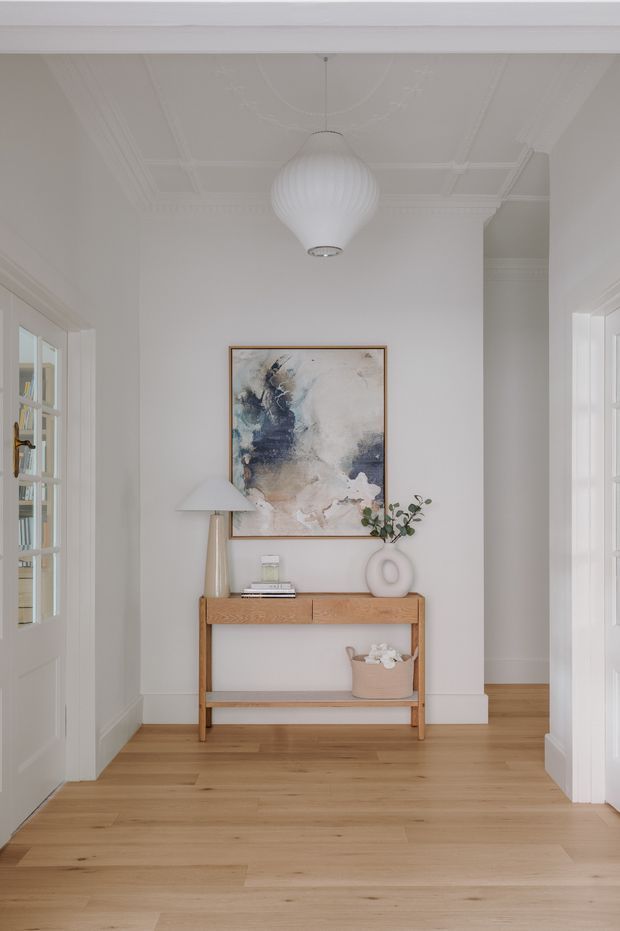 A bright, refined residential hallway or entryway with white painted walls, ornate decorative ceiling mouldings, timber flooring, and white-framed glazed doors. A warm timber console with open shelving sits against the wall beneath an abstract artwork, accompanied by a ceramic lamp and sculptural vase. A ribbed pendant light hangs centrally, creating a calm, softly layered interior.