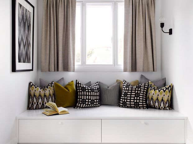 A bright, compact window-seat nook with a white built-in drawer bench beneath a large multi-pane window. Taupe curtains frame the window, while patterned cushions in black, white, grey, mustard, and olive create a graphic accent. A framed monochrome artwork and a simple black wall sconce complete the clean, tailored space.