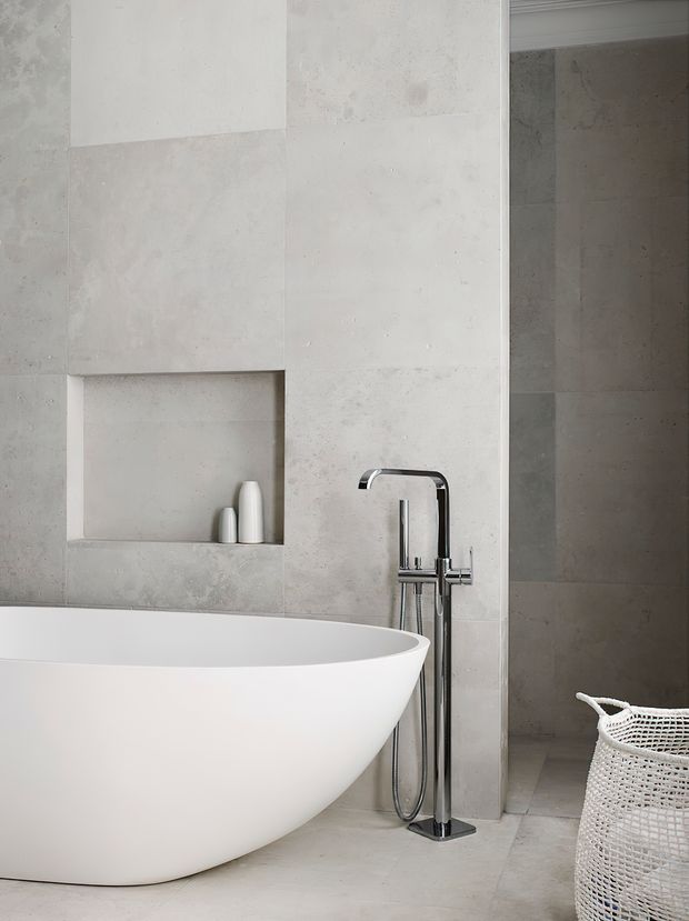 A refined contemporary bathroom featuring a freestanding white oval bathtub, wall-mounted chrome bath filler and handheld shower, large pale grey stone-look wall and floor surfaces, and a recessed display niche with simple white vessels. A woven white basket sits near the shower area, adding a subtle natural texture.