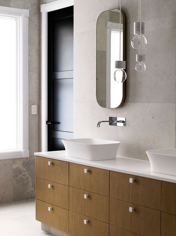 Contemporary bathroom vanity with a long timber-look drawer cabinet, white engineered-stone countertop, two white vessel basins, wall-mounted chrome tapware, and a rounded rectangular mirror. The room has large light grey concrete-look wall tiles, a black internal door, white window trim, and sculptural pendant lights.