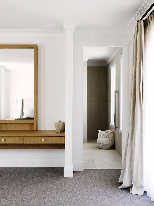 A refined transitional interior showing a carpeted hallway or landing with white walls, decorative moulding and a central structural column. A wall-mounted timber console with integrated drawers and a large timber-framed mirror occupies the left side. Through the doorway is a bright tiled bathroom or ensuite with a freestanding woven basket, windows and a vertical towel rail. Full-height cream curtains frame a bright window on the right.