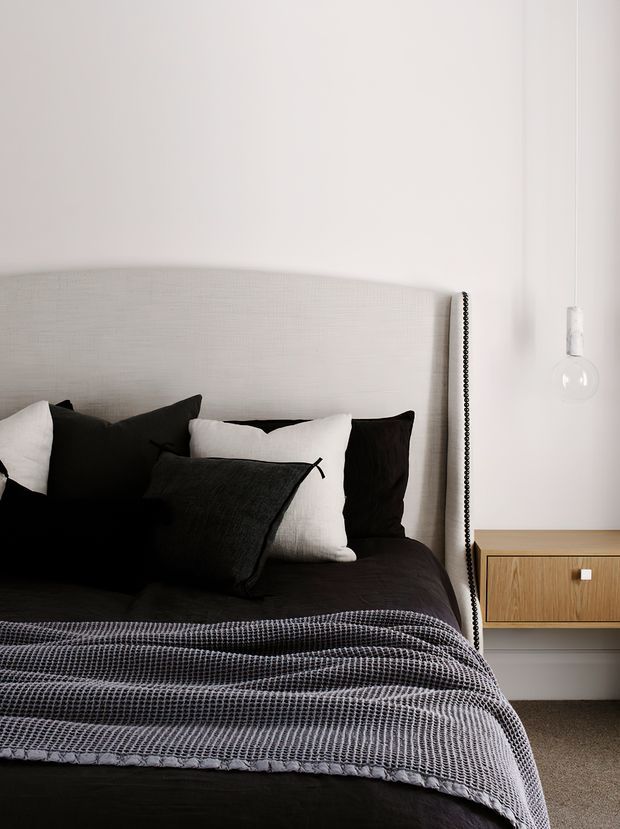 A close-up of a contemporary bedroom featuring a large upholstered light-grey headboard, layered black, white and charcoal bedding, and a simple floating timber bedside drawer with a white knob. A clear pendant bulb hangs beside the bed against plain white walls, creating a restrained monochrome scheme with warm timber detail.