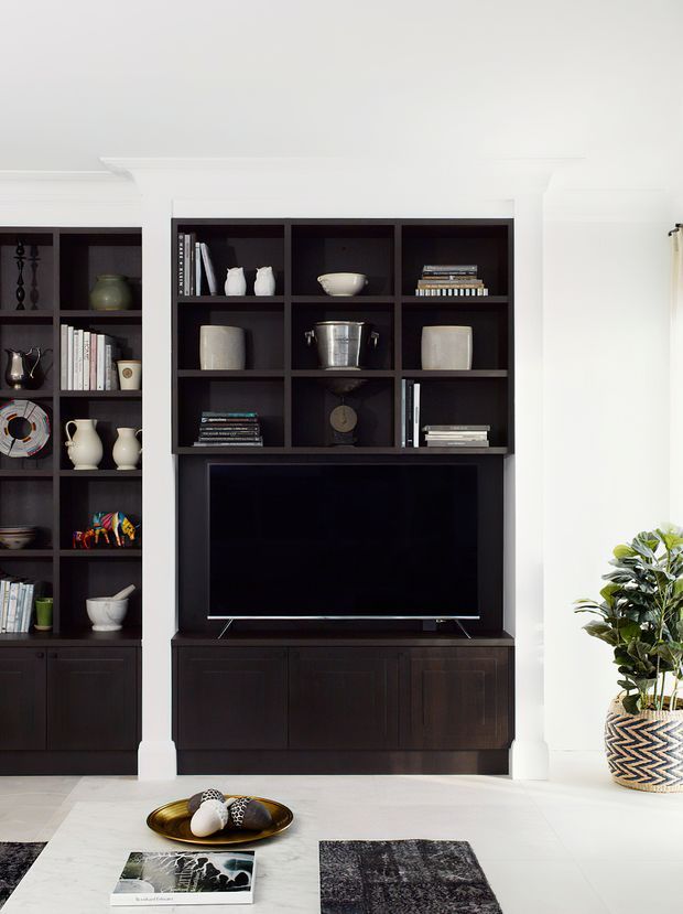 A bright, refined living room wall featuring custom dark built-in cabinetry and open shelving surrounding a recessed flat-screen television. The shelving displays books, ceramics, decorative objects, and tableware, while a large potted plant, coffee table, rugs, and white architectural trim complete the room.
