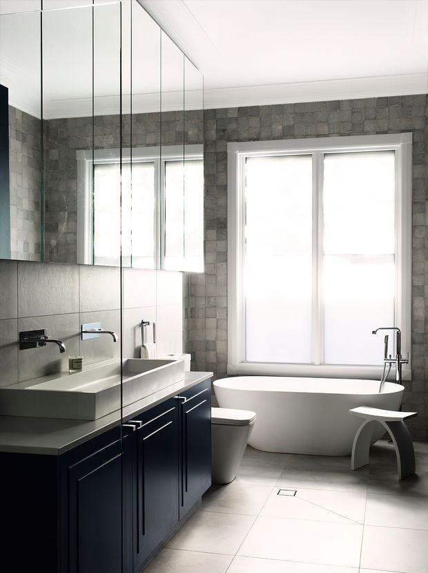 A bright, refined bathroom with a freestanding white bathtub beneath two tall windows, a wall-mounted faucet, double vanity with dark navy cabinetry and pale benchtop, a toilet, large-format light floor tiles, and grey textured wall tiles. Full-height mirrored cabinetry or wall panels reflect the vanity and windows, while a small sculptural stool sits beside the tub.