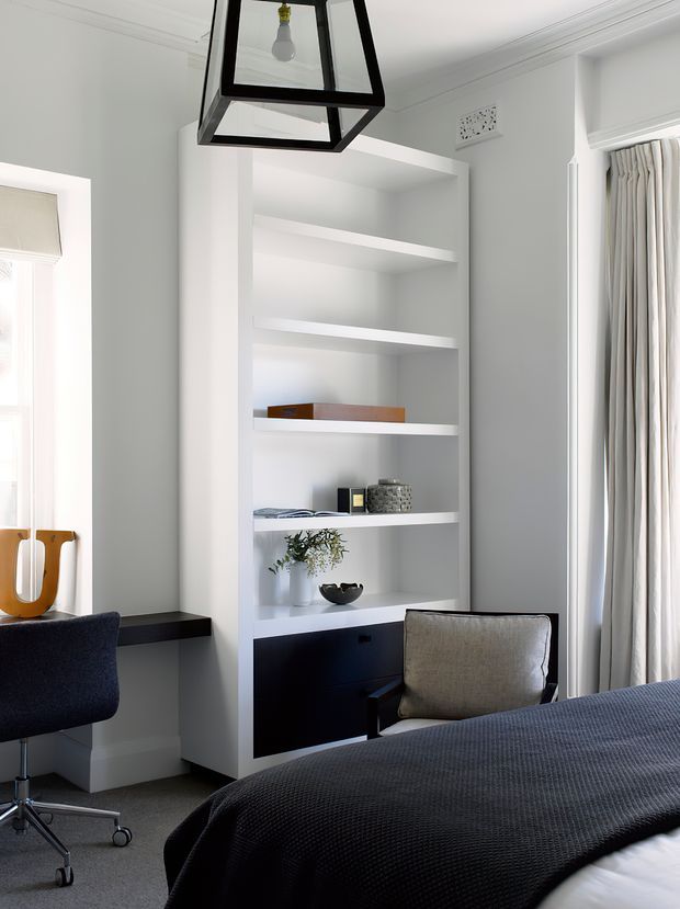 Bright, refined bedroom interior with a built-in white open shelving unit and dark lower cabinetry, a partially visible bed dressed in dark textured bedding, an upholstered armchair, and a compact desk chair by the window. Full-height curtains, pale walls with decorative ceiling moulding, and a black geometric pendant light create a clean contemporary setting with contrasting dark accents.