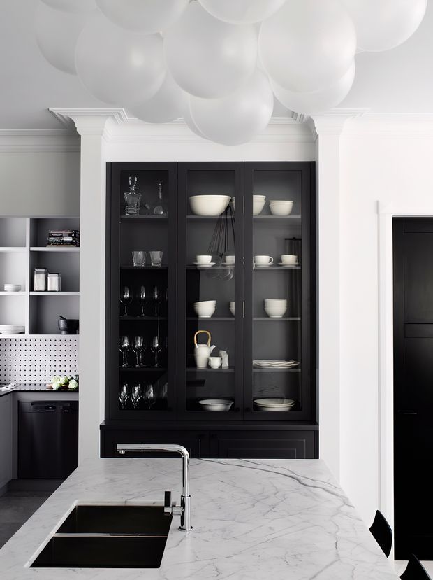 A refined kitchen interior featuring a large white marble island with an undermount sink and chrome tap, dark cabinetry, open shelving, and a tall black glass-front display cabinet filled with white tableware and glassware. White paneled walls and ornate cornice detailing contrast with the black joinery, while oversized white spherical pendant forms create a dramatic ceiling installation. A black-framed opening is visible at right.