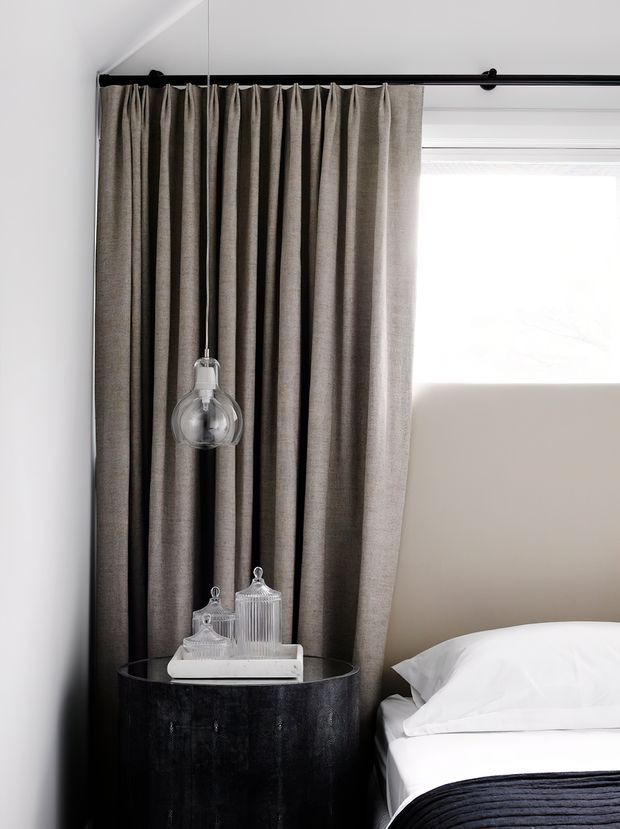 A refined bedroom corner featuring a bed with crisp white linens and a dark charcoal throw, a round black bedside table with decorative glass vessels, and a clear globe pendant light. Full-height taupe-grey curtains frame a bright window, creating a subdued, elegant atmosphere with strong vertical drapery and dark accents.