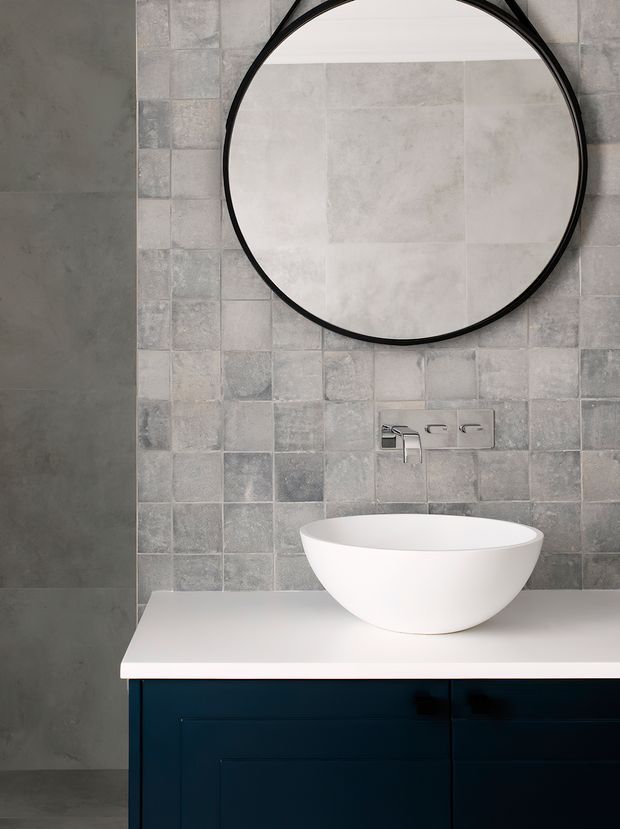 A contemporary bathroom vanity featuring a white vessel basin on a white benchtop, dark navy-blue cabinetry, a chrome wall-mounted tap, and a large round black-framed mirror. The walls are clad in varied grey square tiles, with darker grey concrete-look surfaces at the side and floor.