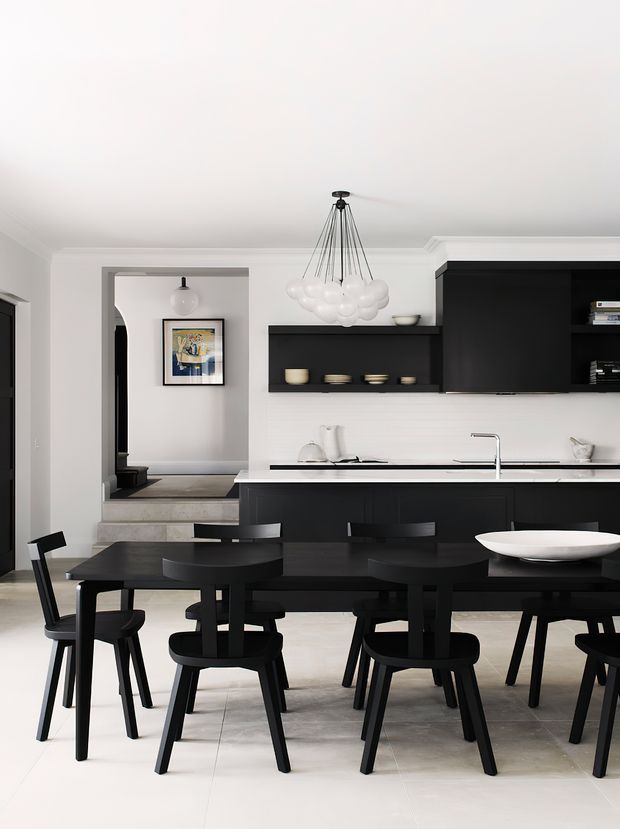 A refined open-plan dining and kitchen interior with a predominantly black-and-white palette. A large black dining table with matching chairs occupies the foreground, while a black island and dark built-in kitchen cabinetry sit against white tiled walls. The room features pale stone or tile flooring, white walls with decorative cornices, a sculptural white pendant light, minimalist artwork, and a small step-up entry toward an adjacent area.