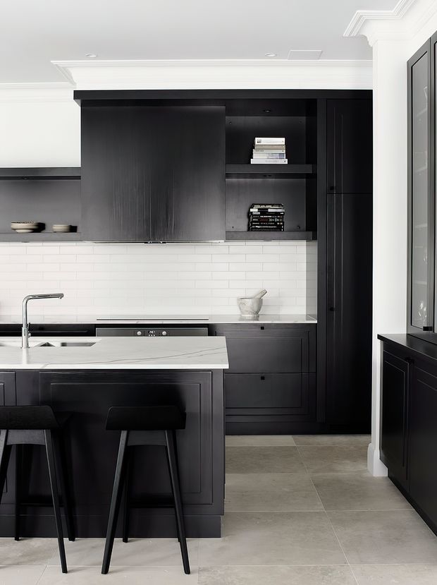 A refined, high-contrast contemporary kitchen with matte black built-in cabinetry, open shelving, white subway-tile splashback, and a light veined stone island benchtop. Black stools sit at the island, while polished white ceiling mouldings and neutral large-format floor tiles add traditional detailing and balance the dark joinery.