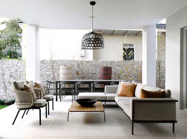 Bright, covered outdoor living and dining area with a contemporary white-lined ceiling, pale tiled floor, stone boundary wall, and open garden outlook. The space contains a large L-shaped sofa, woven lounge chairs, a low timber coffee table, dining table and chairs, and a distinctive black woven pendant light. A sheltered patio structure and mature planting create a refined indoor-outdoor entertaining setting.