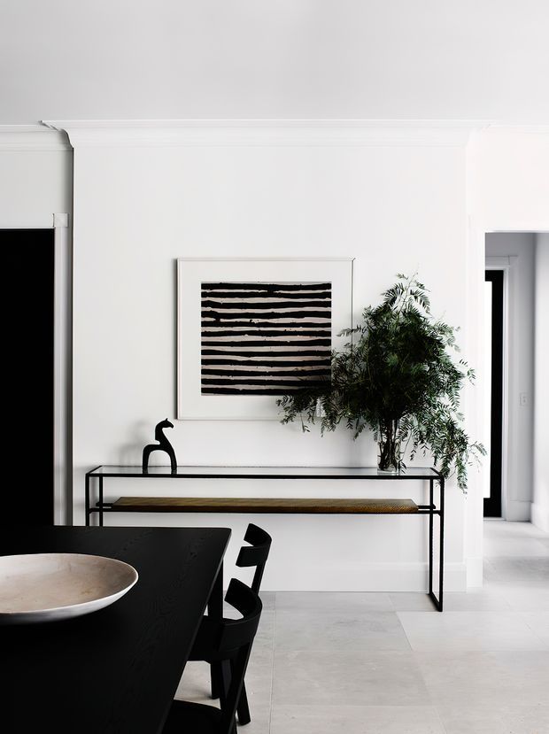 Bright, refined interior view of a dining and entry area. A black dining table and chairs occupy the foreground, while a slim black metal console with a timber bench sits against a white wall beneath abstract striped artwork. A sculptural black vase and leafy indoor tree add contrast. Pale large-format floor tiles, white walls, simple cornice detailing, and dark door openings create a clean contemporary composition.