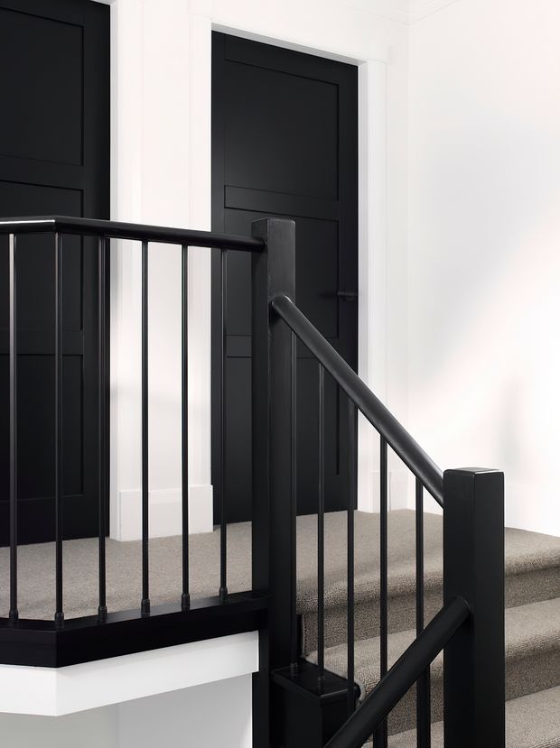 A bright, minimalist stairway and landing with black painted timber panelled doors, white walls and trim, grey carpeted stairs, and a black metal balustrade with square newel posts and slender vertical spindles.