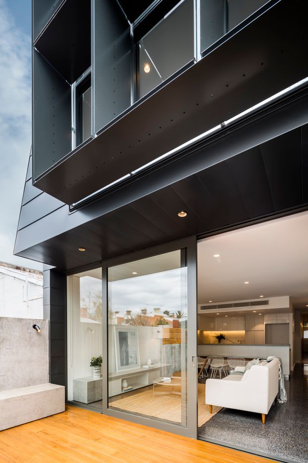 A contemporary multi-level residential extension viewed from an outdoor deck, with dark metal cladding, projecting upper-floor balcony, large glazed sliding doors, and recessed exterior lighting. Through the glazing, a bright open-plan living, dining and kitchen area is visible, featuring light cabinetry, a dining table, white sofas, timber flooring, and a dark speckled floor finish.