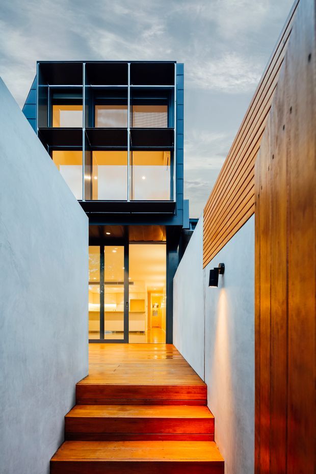 A narrow exterior passage and stairway leading up to a modern multi-level residence. Warm timber steps and decking contrast with smooth white rendered walls, a tall timber-clad boundary wall, and a projecting upper volume with dark metal framing and extensive glazing. Warm interior lighting is visible through the glass, with discreet wall-mounted exterior lighting along the passage.