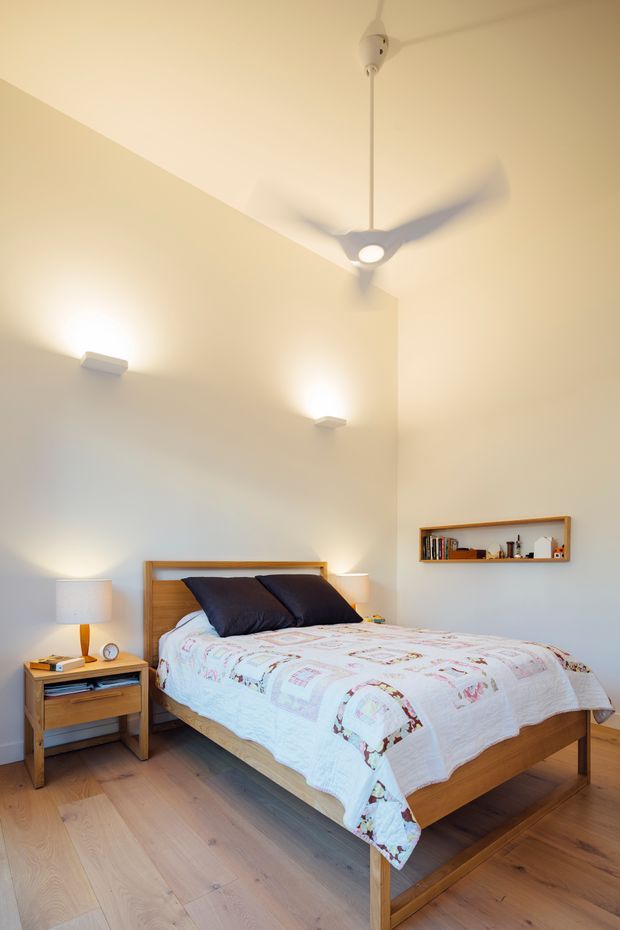 Bright, airy bedroom with a timber bed and matching bedside table on a light timber floor. The room has pale walls, a high sloping ceiling, a suspended ceiling fan with integrated light, wall sconces, a floating timber shelf, and a colourful patterned quilt with dark pillows.