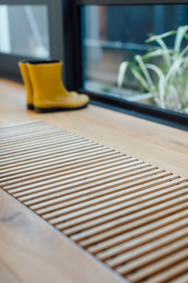 Close-up interior detail of a pale timber slatted floor grate or raised mat beside a large dark-framed window. A bright yellow rubber boot rests on the timber floor, with a leafy plant visible beyond the glazing. The image has a warm, natural, contemporary feel and focuses on the floor detailing and window junction.