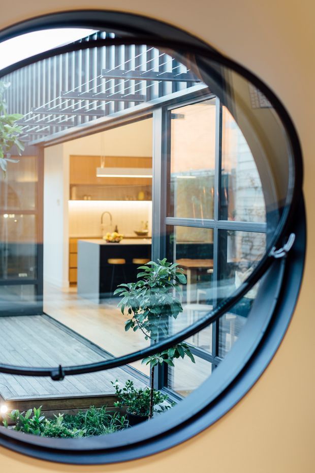 View through a large circular black-framed window into a contemporary courtyard-style home. The image shows a timber deck, extensive glazed doors and windows, a planted interior courtyard, and a warm modern kitchen with timber cabinetry, a dark island and integrated lighting. A glazed or louvred overhead structure provides filtered daylight.