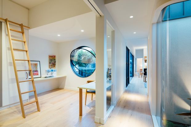 Bright contemporary residential interior showing a compact study or work nook opening onto a long hallway. The space has white walls and ceilings, pale timber flooring, built-in white shelving, a timber ladder, framed artwork, a small desk, and distinctive round glazing. The hallway extends toward additional rooms, with blue-tinted glazing and textured glass doors on the right.