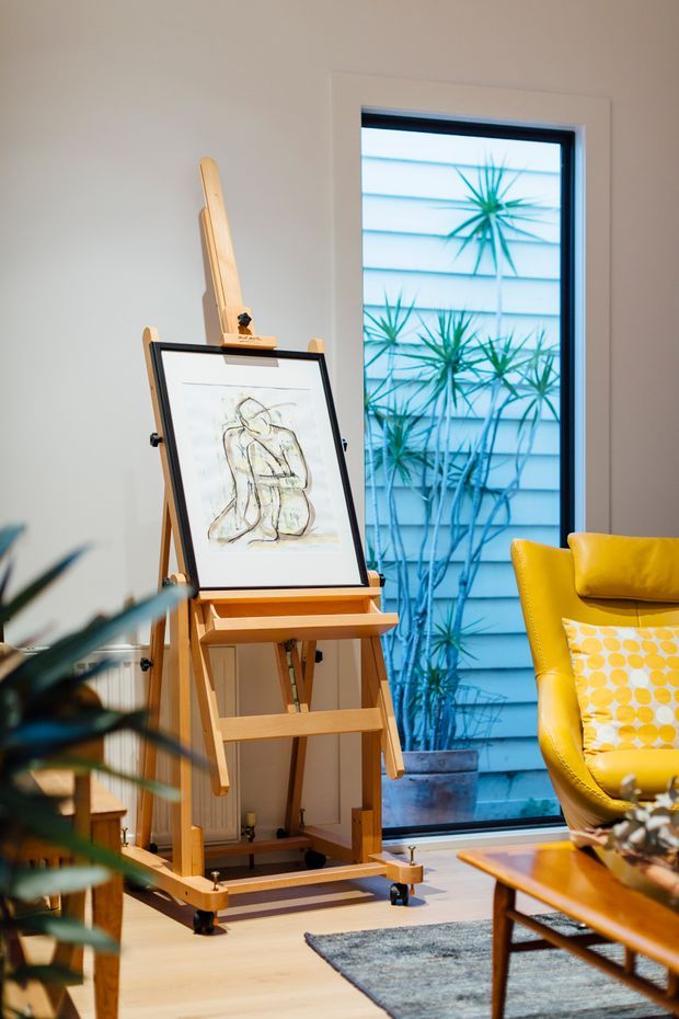 A warmly lit living room corner featuring a timber artist's easel with framed artwork, a vivid yellow upholstered lounge chair, indoor plants, a timber coffee table, and a tall window overlooking exterior planting and cladding. The space has light timber flooring and a relaxed, art-focused character.