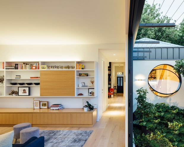 A bright contemporary living room with built-in white shelving and timber cabinetry, opening through full-height glazing to a landscaped courtyard. The room includes a low media/storage wall, pale timber flooring, a textured rug, and upholstered seating. Outside, a circular feature window and grey metal roof are visible on an adjoining structure.