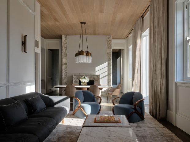 A refined open-plan living and dining room with a dark curved sectional sofa, blue upholstered lounge chairs, and a marble coffee table in the foreground. The room has light decorative wall panelling, tall windows with full-length curtains, a pale timber-clad ceiling, and a dining table positioned near a marble fireplace surround. A sculptural pendant light hangs centrally over the dining area, creating a bright, elegant interior with strong natural light.