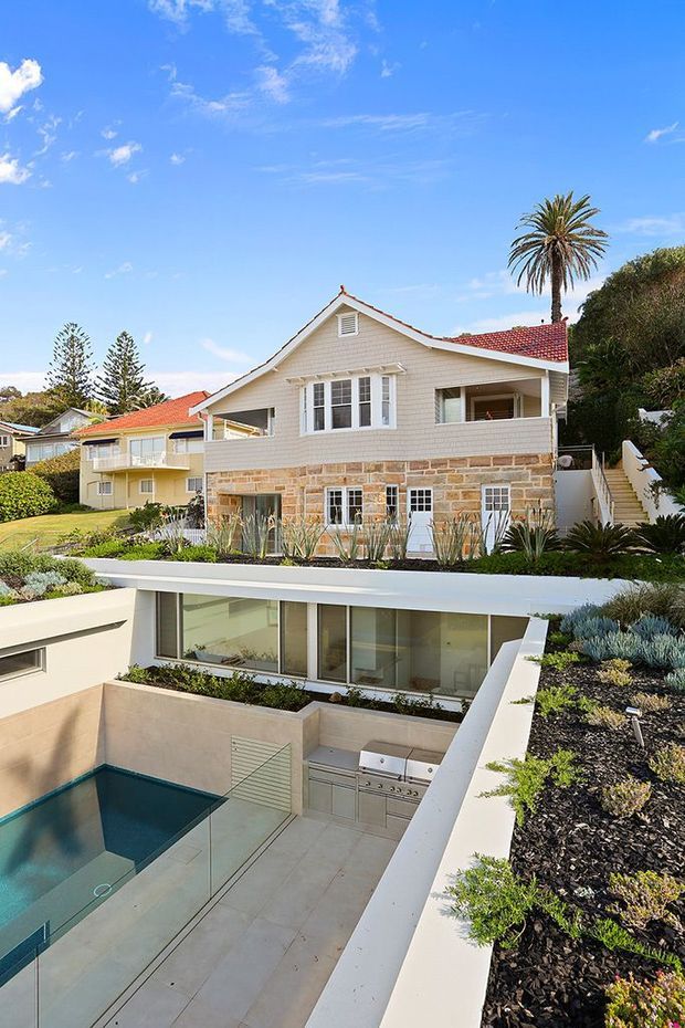 A bright exterior view of a multi-level coastal-style home with a cream weatherboard upper storey, sandstone masonry lower level, and red tiled pitched roof. The property features a landscaped terrace, built-in outdoor barbecue, large glazed lower-level room, and a sunken swimming pool with glass balustrading. Mature palms and neighbouring homes are visible under a clear blue sky.