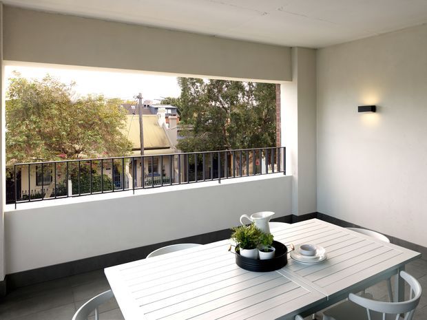Covered outdoor dining terrace or balcony with a white slatted dining table, curved white chairs, a black metal balustrade, pale rendered walls, and a compact wall light. The open side overlooks neighbouring rooftops and trees. The space has a clean, contemporary feel with dark floor and trim accents.