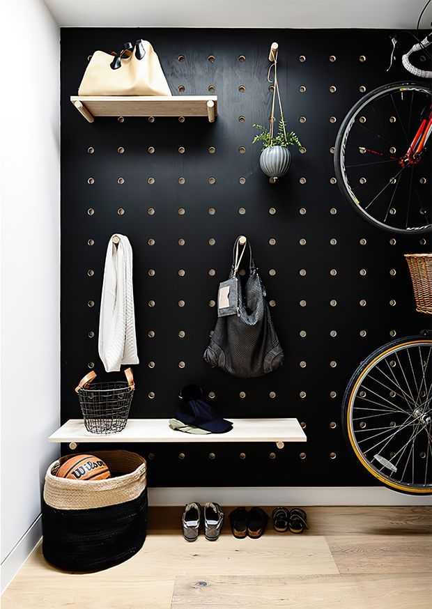 A compact, highly organised entryway or mudroom storage wall with a matte black perforated pegboard, timber shelves, hooks, and integrated display storage. Bicycles, bags, hats, shoes, a towel, basket and hanging planter are arranged on the wall, with pale timber flooring and white surrounding walls creating strong contrast.