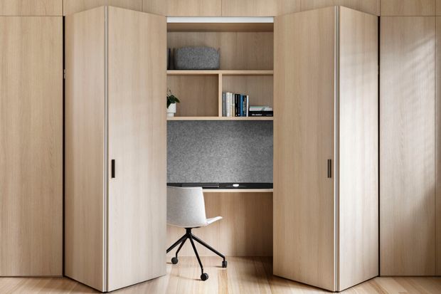 A compact built-in timber study nook concealed behind tall bi-fold or pivoting cabinetry doors. The open workspace includes a desk, shelving with books and decor, a dark grey acoustic or textured backing panel, and a white swivel office chair. Light timber flooring and matching wood cabinetry create a warm, seamless fitted interior.
