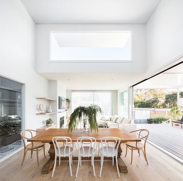 Bright open-plan dining and living area with a large timber dining table, mixed timber and white dining chairs, pale timber flooring, white walls, and extensive glazing opening onto a covered timber deck. A high-level window and open ceiling volume create an airy, light-filled interior, with built-in shelving and a fireplace visible to the side.