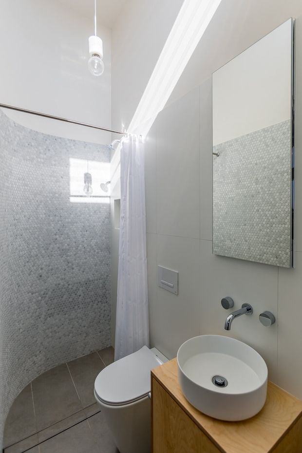 Bright contemporary bathroom with a walk-in shower enclosed by a white curtain, pale small-format mosaic wall tiles, a wall-mounted mirror, floating wall-mounted tapware, a round vessel basin, and a timber vanity. The room has dark grey tiled flooring, white walls, and a minimalist compact layout.