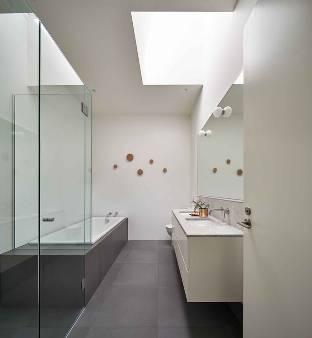 Contemporary minimalist bathroom with a freestanding rectangular bathtub, frameless glass shower enclosure, wall-mounted vanity with double basins, large mirror, and dark large-format tiled flooring. White walls and ceiling are punctuated by a large skylight, while small round timber-toned wall accents add warmth.