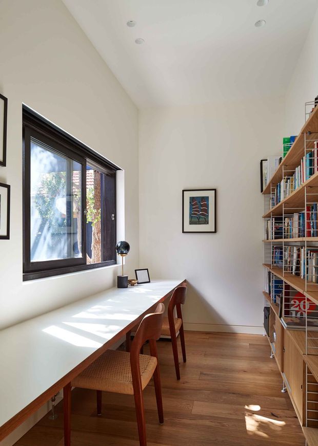 A bright, narrow home office or study with a long built-in timber desk positioned beneath a dark-framed window. Two wooden chairs, a small desk lamp, framed artwork, and extensive open timber bookshelves filled with books line the room. The space has white walls, recessed ceiling lights, and warm timber flooring, with strong natural sunlight entering through the window.
