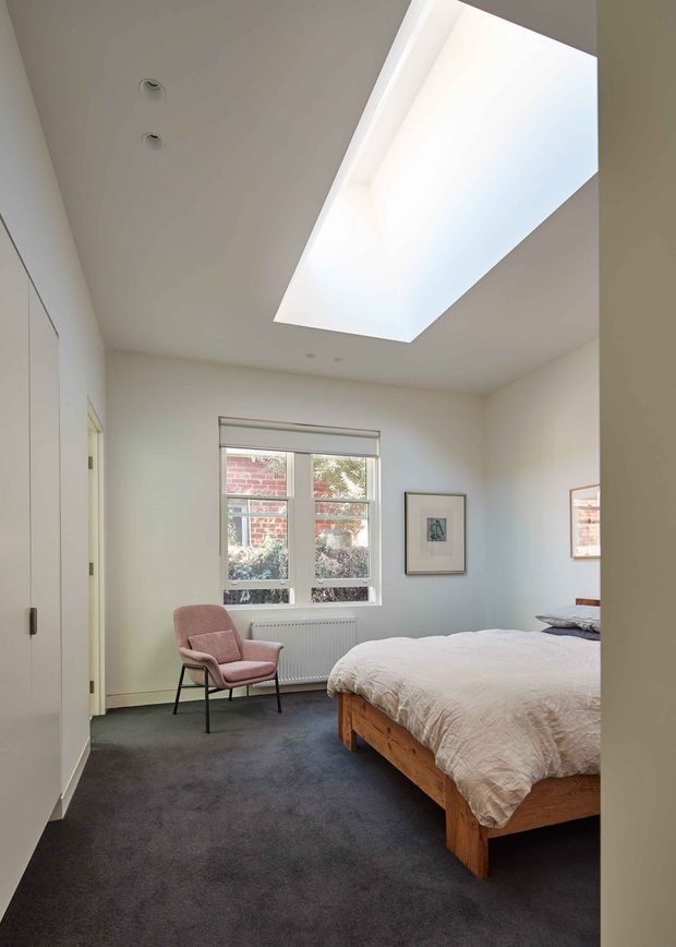 Bright contemporary bedroom viewed from the doorway, featuring a large rectangular skylight, white walls and ceiling, dark charcoal carpet, a timber bed with light bedding, a blush upholstered lounge chair, built-in white wardrobe cabinetry, framed artwork, and a window overlooking brickwork and planting. The room has a calm, simple, natural-light-filled atmosphere.