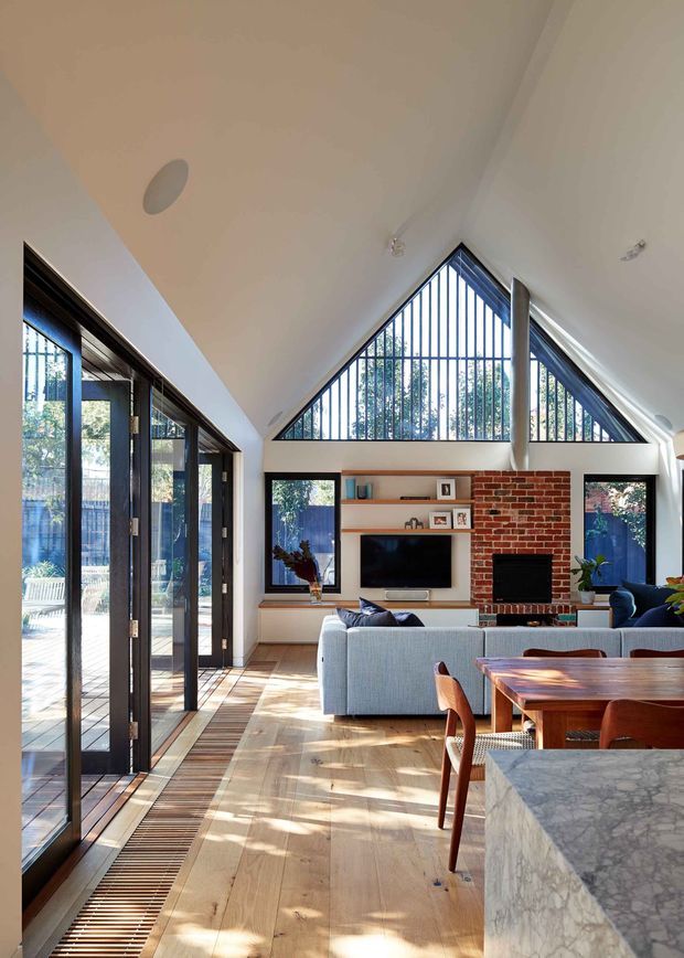 Bright open-plan living and dining area with a dramatic vaulted ceiling and triangular clerestory glazing fitted with vertical security bars. Full-height black-framed glazed folding doors open to an outdoor area. The room features pale timber flooring, a red exposed-brick fireplace wall with integrated shelving and television, a light grey sofa, and a substantial marble kitchen island in the foreground.
