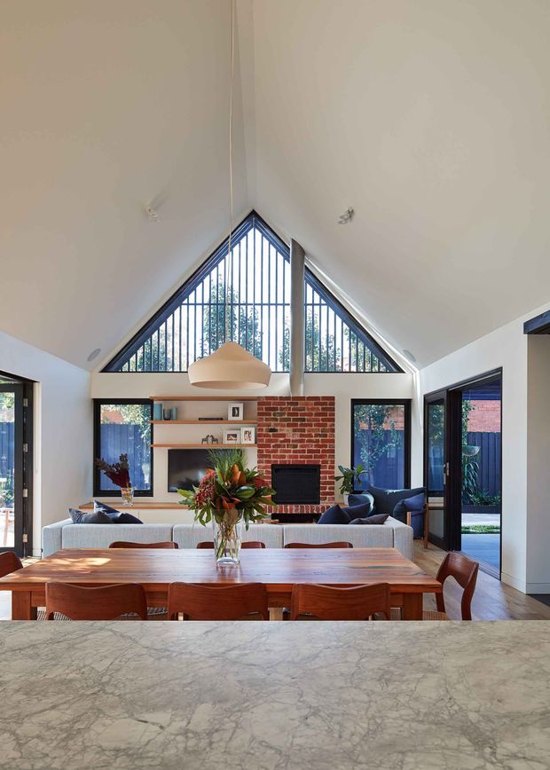 A bright open-plan living and dining area viewed from behind a stone kitchen benchtop. The room features a dramatic vaulted ceiling with a triangular glazed gable and dark vertical window bars, exposed red brick around a fireplace, built-in timber shelving, a large timber dining table, upholstered seating, and extensive black-framed glazing opening toward an outdoor courtyard.