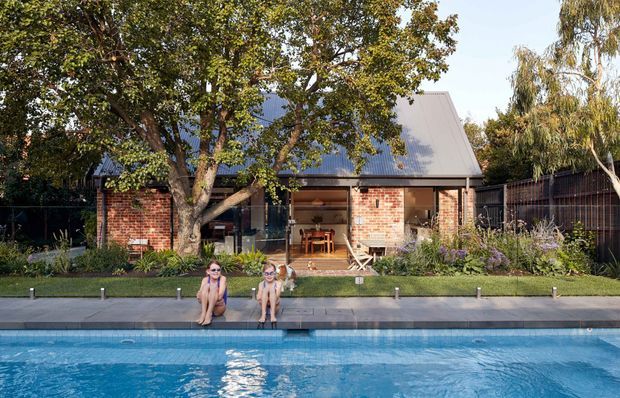 A single-storey brick residential building with a steep pitched metal roof, broad glazed opening, and sheltered outdoor dining area, viewed across a swimming pool. Mature trees, planted garden beds, fencing, and a paved pool edge surround the house.