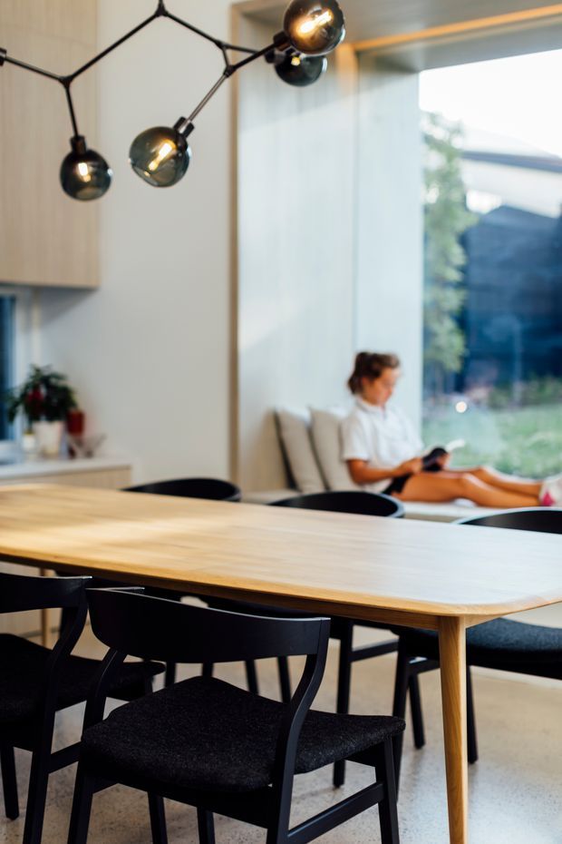 Contemporary dining area with a large light timber dining table surrounded by black upholstered dining chairs. A sculptural black multi-arm pendant light hangs overhead, while pale built-in cabinetry and a full-height glazed opening bring soft natural light into the room. A person is seated in the adjacent lounge area in the background.